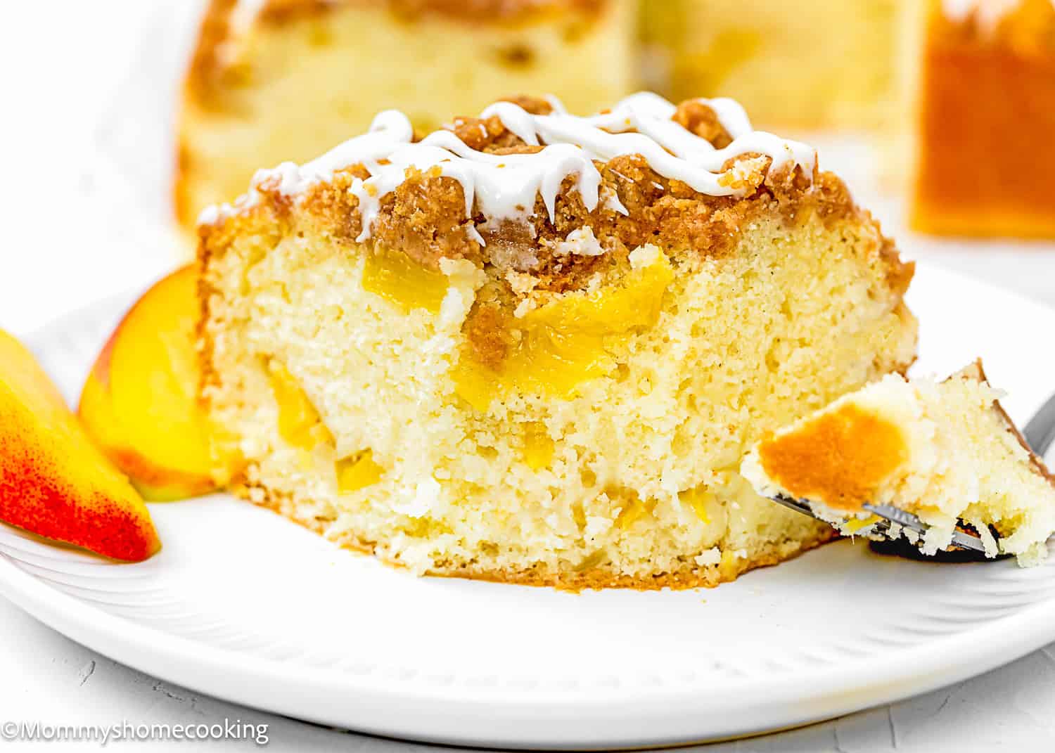 A slice of Eggless Peach Coffee Cake with crumb topping and white icing sits on a white plate, accompanied by peach slices and a fork holding a bite-sized piece.