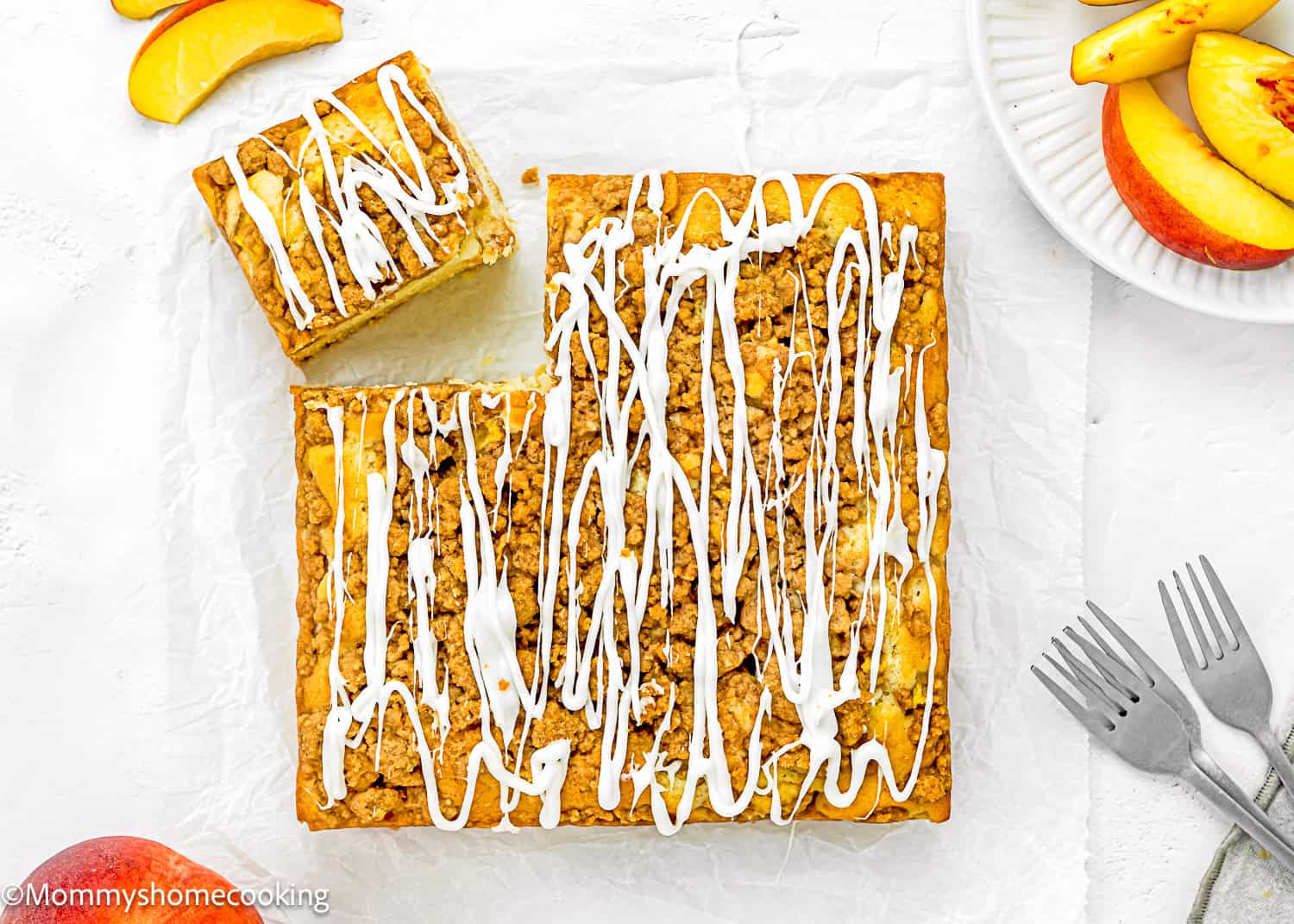 A rectangular Eggless Peach Coffee Cake with white icing drizzle, some pieces cut, on parchment paper. Sliced peaches and forks are nearby on a white surface.