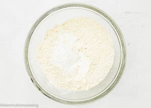 A clear glass bowl containing a mixture of dry ingredients, including flour and baking powder, for an Eggless Peach Coffee Cake, on a white surface.