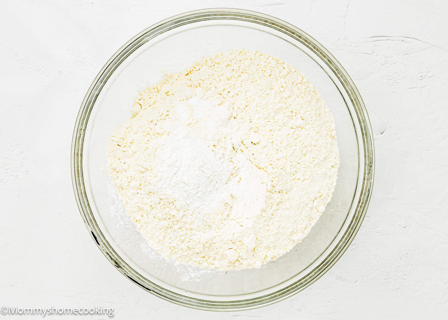A clear glass bowl containing a mixture of dry ingredients, including flour and baking powder, for an Eggless Peach Coffee Cake, on a white surface.