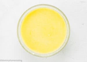 A clear glass bowl filled with a yellow, frothy liquid mixture for Eggless Peach Coffee Cake sits on a white textured surface.