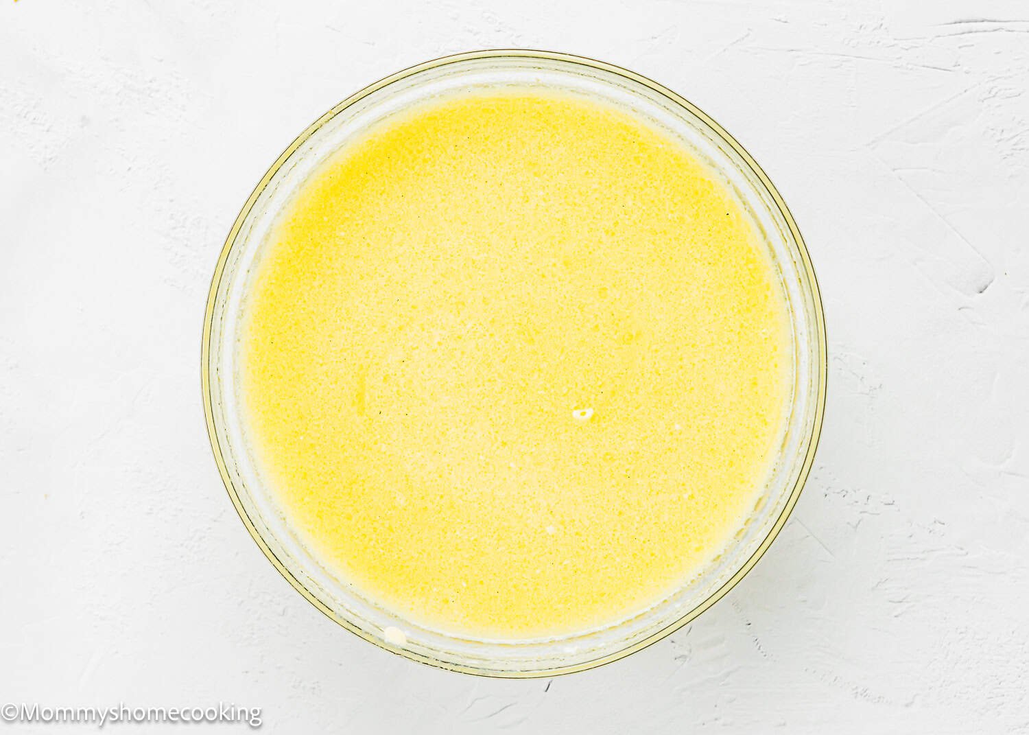 A clear glass bowl filled with wet ingredients mixture for Eggless Peach Coffee Cake sits on a white textured surface.