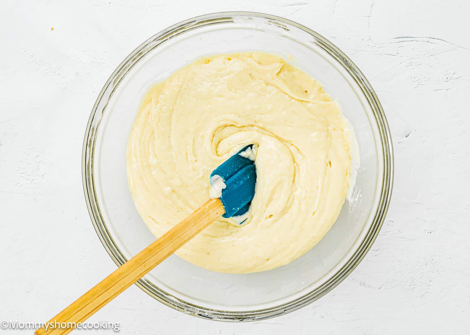 A glass bowl with creamy batter for Eggless Peach Coffee Cake being mixed with a blue silicone spatula on a white surface.