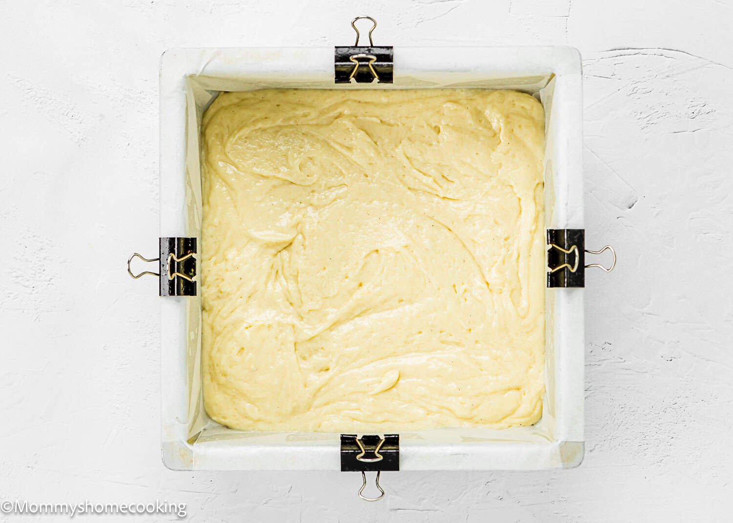 A square baking pan lined with parchment paper and binder clips holds an even layer of Eggless Peach Coffee Cake batter, ready to be baked.