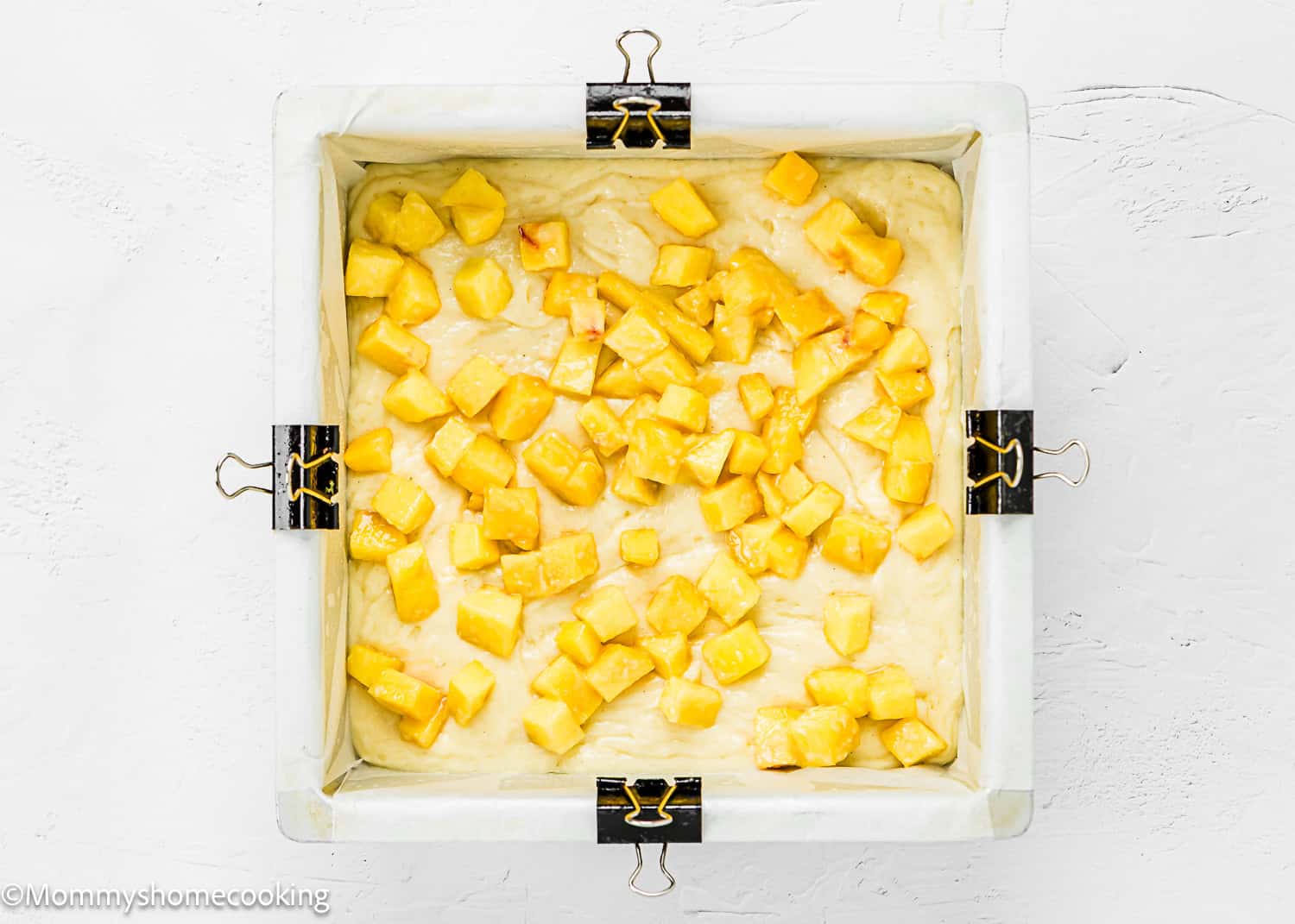Square baking pan lined with parchment paper, filled with Eggless Peach Coffee Cake batter and topped with chopped peach pieces, held in place with binder clips.