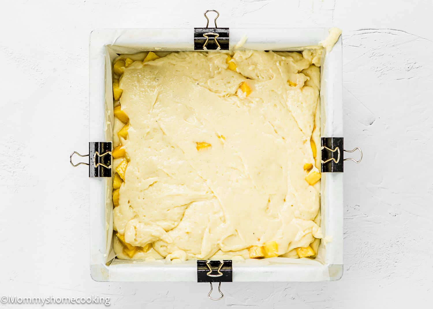 A square baking pan lined with parchment paper and binder clips, filled with unbaked Eggless Peach Coffee Cake batter containing visible fruit pieces.