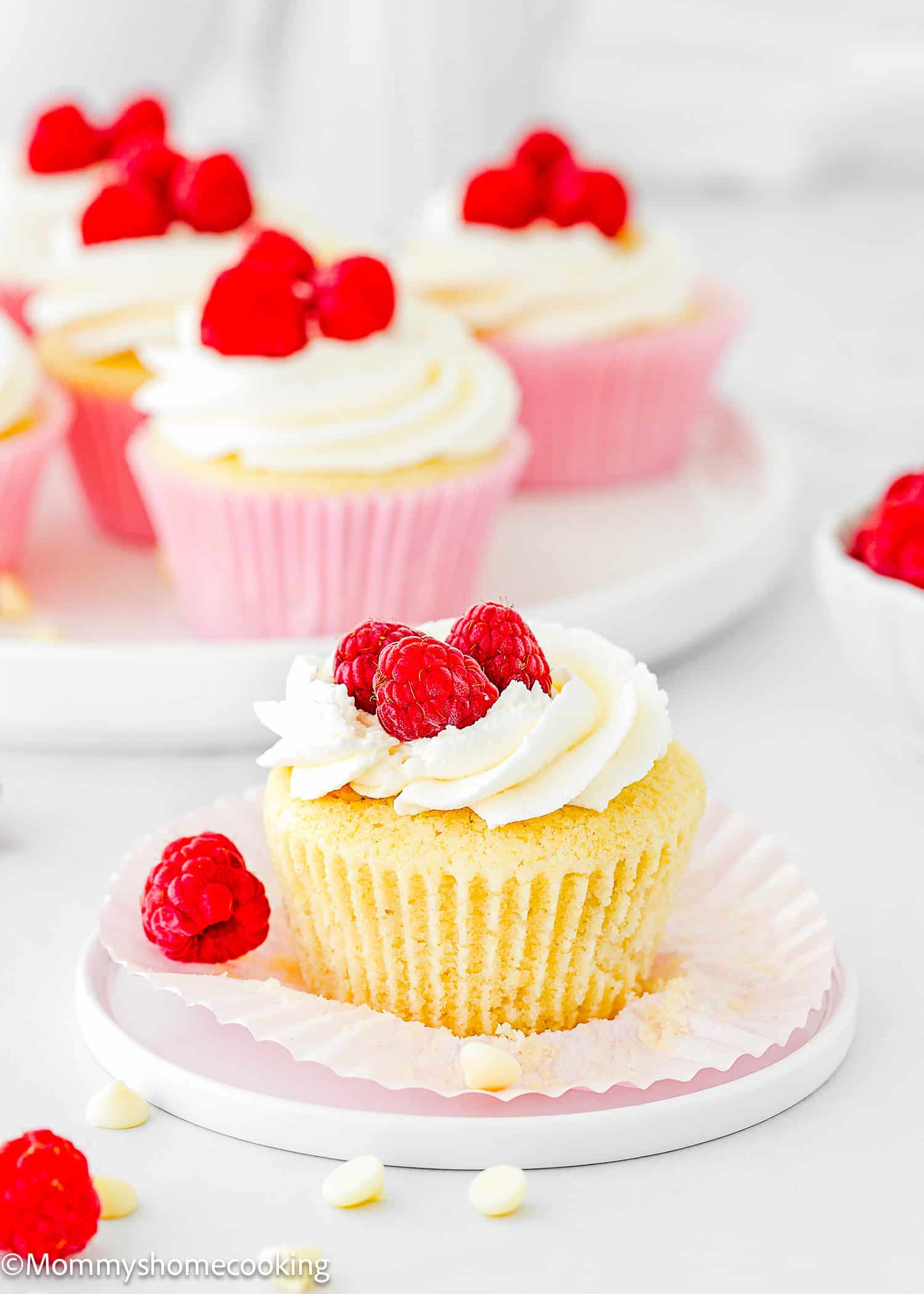 An Eggless White Chocolate Raspberry Cupcake with white frosting and fresh raspberries on top sits on a plate with its wrapper partially unwrapped; more cupcakes and raspberries are in the background.