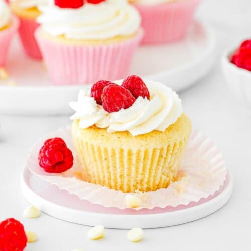 An Eggless White Chocolate Raspberry Cupcake with white frosting and fresh raspberries on top sits on a plate with its wrapper partially unwrapped; more cupcakes and raspberries are in the background.