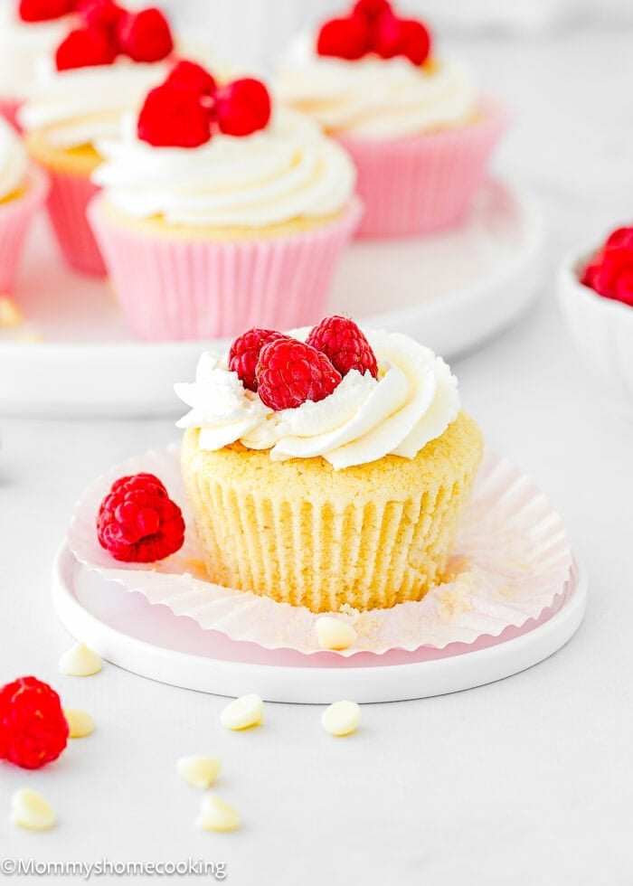 An Eggless White Chocolate Raspberry Cupcake with white frosting and fresh raspberries on top sits on a plate with its wrapper partially unwrapped; more cupcakes and raspberries are in the background.