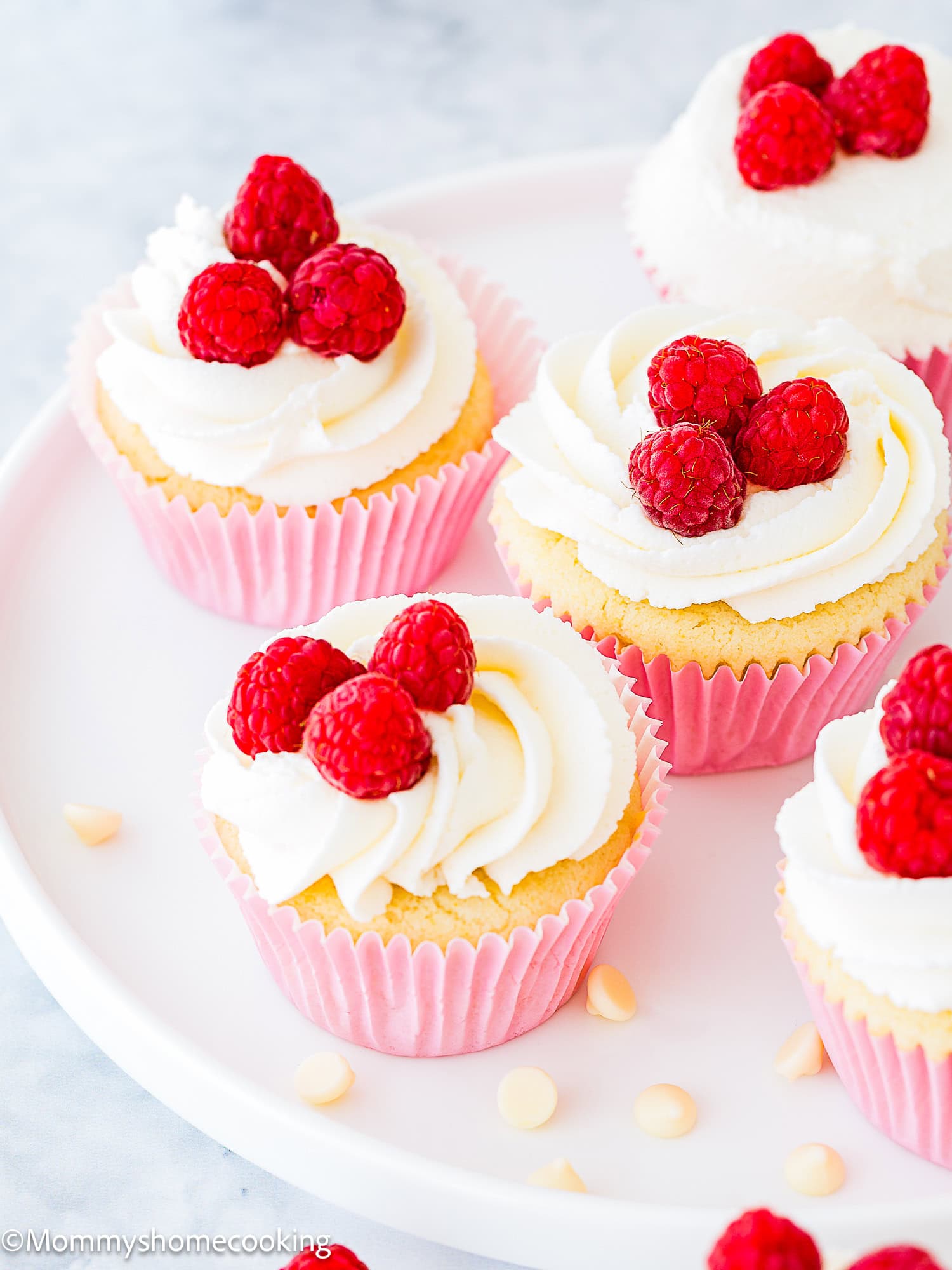 Eggless White Chocolate Raspberry Cupcakes with white frosting, topped with fresh raspberries and a sprinkling of white chocolate chips, arranged on a white plate.