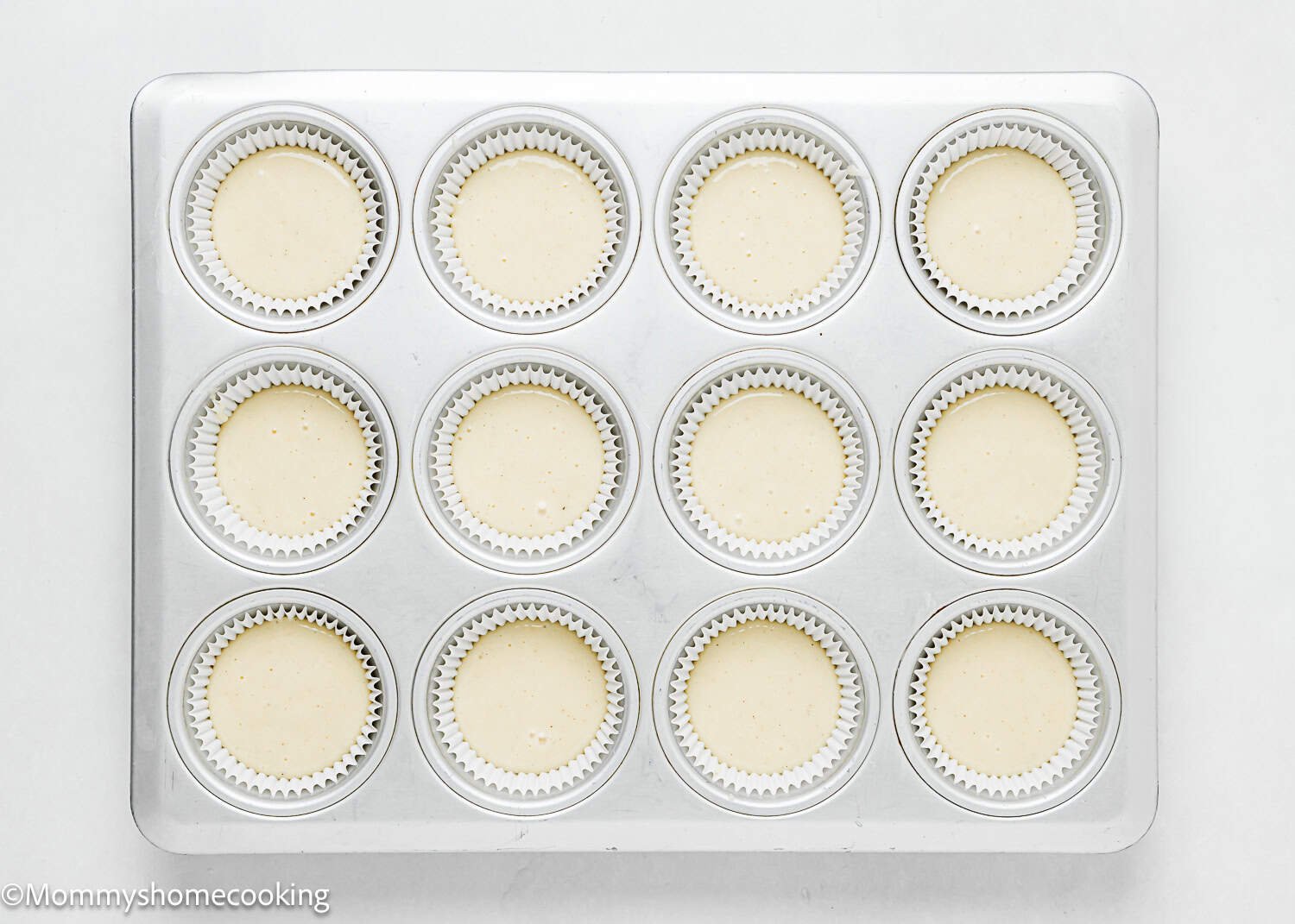 A muffin tin filled with twelve cupcake liners, each containing unbaked Eggless White Chocolate Raspberry Cupcake batter, viewed from above on a white surface.