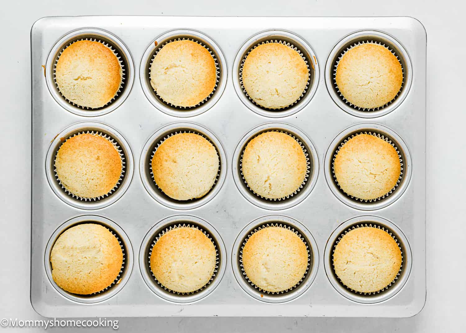 A metal muffin tray with twelve freshly baked Eggless White Chocolate Raspberry Cupcakes in paper liners, viewed from above on a white surface.
