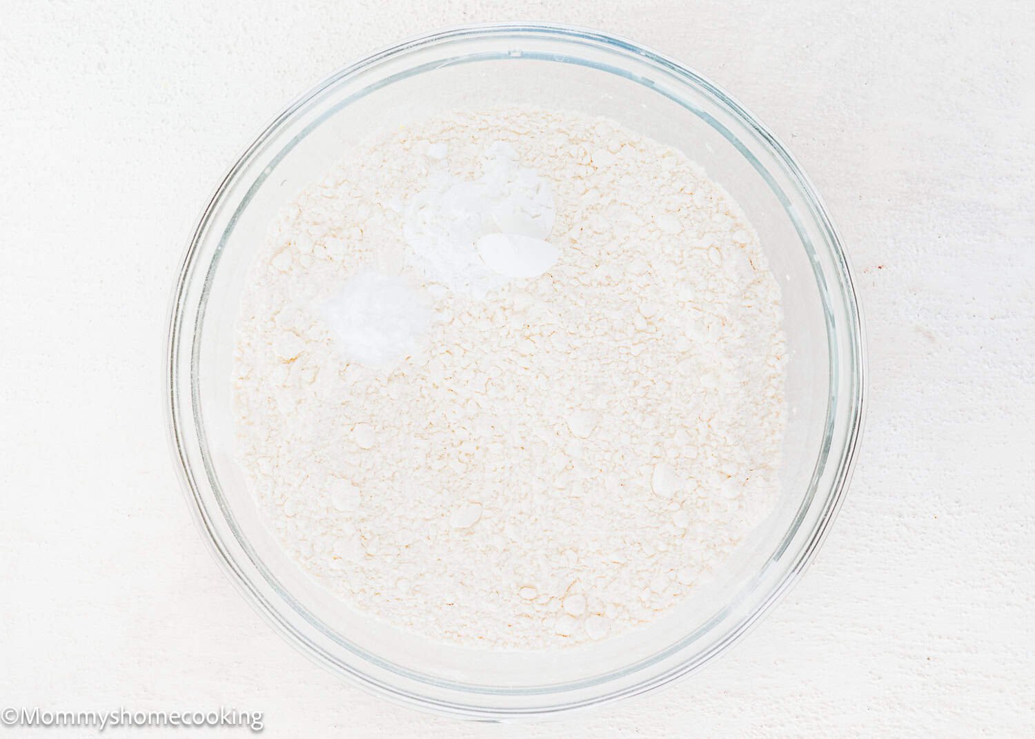 A glass bowl containing dry flour mixture with some white powder, possibly baking soda or baking powder, for Eggless White Chocolate Raspberry Cupcakes, on a white surface.