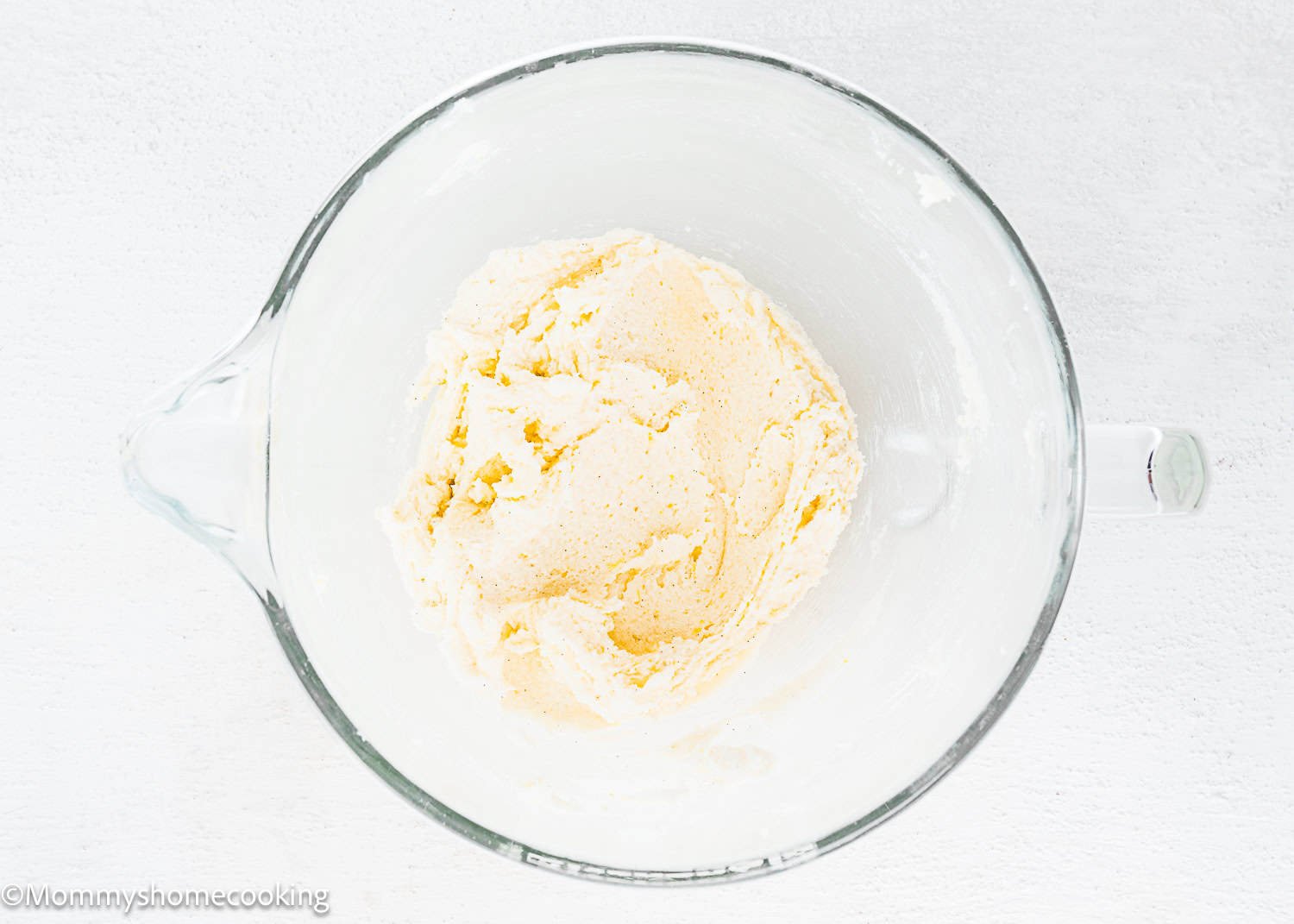 A glass mixing bowl containing a pale, creamy mixture for Eggless White Chocolate Raspberry Cupcakes sits on a white surface.