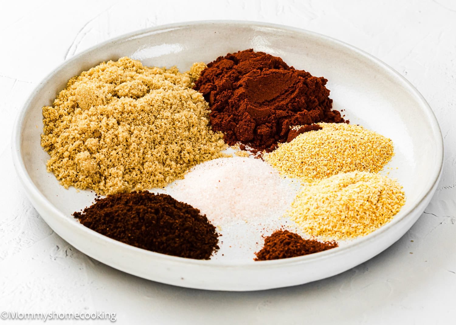 A white plate displays separate piles of brown, reddish-brown, yellow, and white powdered spices and seasonings—perfect for creating a homemade barbecue meat rub.