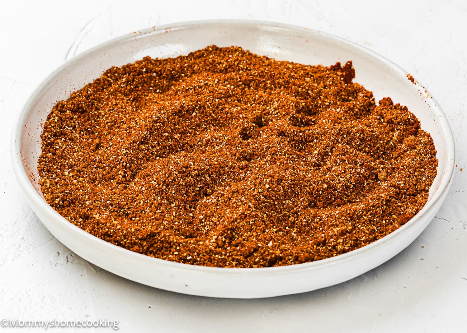 A white plate filled with a coarse, reddish-brown Homemade Barbecue Meat Rub sits on a white surface.