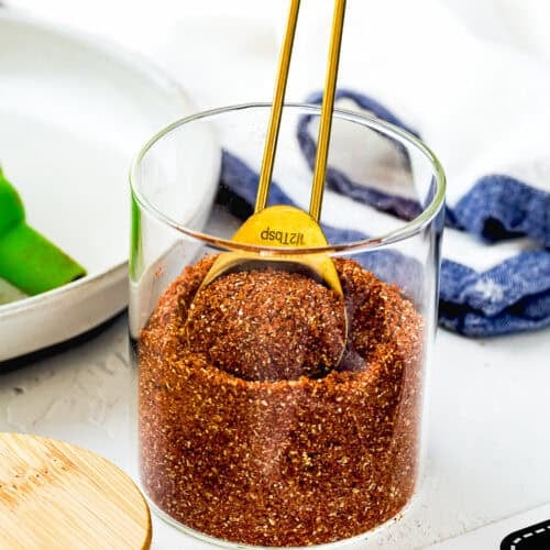 A glass jar filled with Homemade Barbecue Meat Rub and a gold measuring spoon, placed on a white surface with a wooden lid and label nearby.