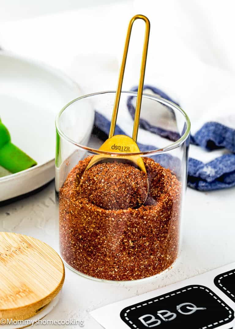 A glass jar filled with Homemade Barbecue Meat Rub and a gold measuring spoon, placed on a white surface with a wooden lid and label nearby.