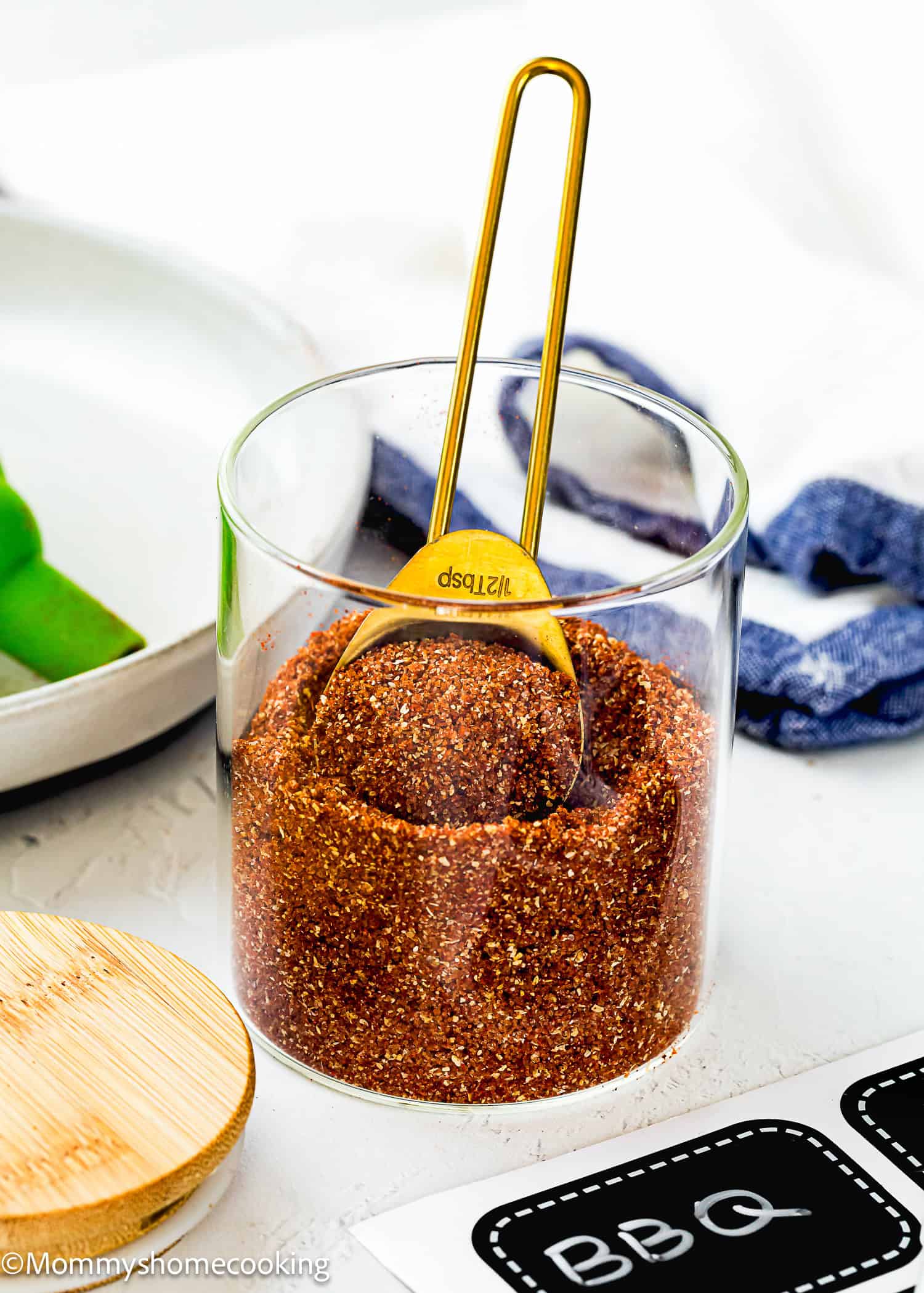 A glass jar filled with Homemade Barbecue Meat Rub and a gold measuring spoon, placed on a white surface with a wooden lid and label nearby.