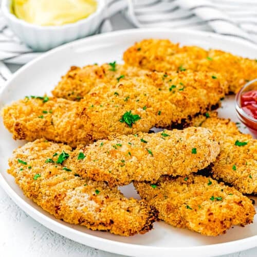 A white plate with several Homemade Chicken Tenders (Egg & Dairy-Free), breaded and baked, garnished with chopped parsley, served with ketchup and a bowl of yellow dipping sauce in the background.