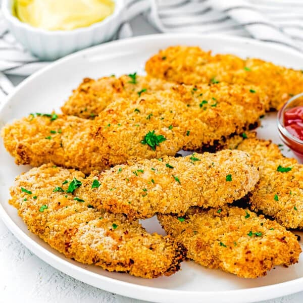 A white plate with several Homemade Chicken Tenders (Egg & Dairy-Free), breaded and baked, garnished with chopped parsley, served with ketchup and a bowl of yellow dipping sauce in the background.