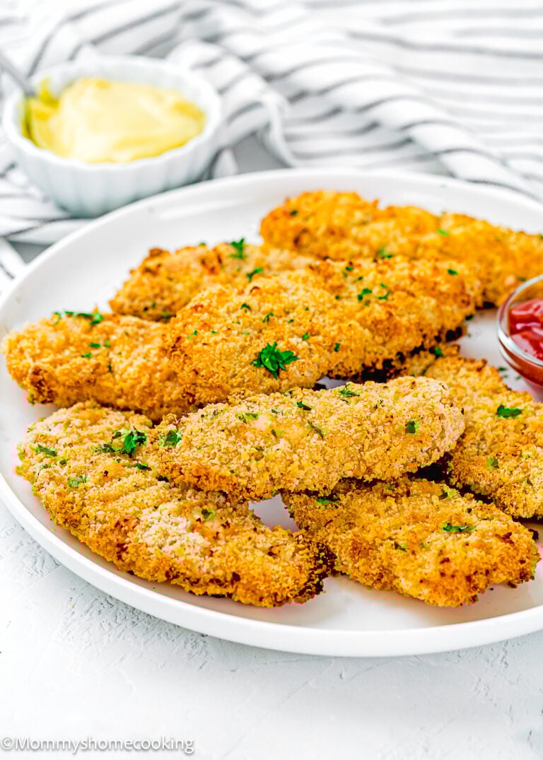 A white plate with several Homemade Chicken Tenders (Egg & Dairy-Free), breaded and baked, garnished with chopped parsley, served with ketchup and a bowl of yellow dipping sauce in the background.