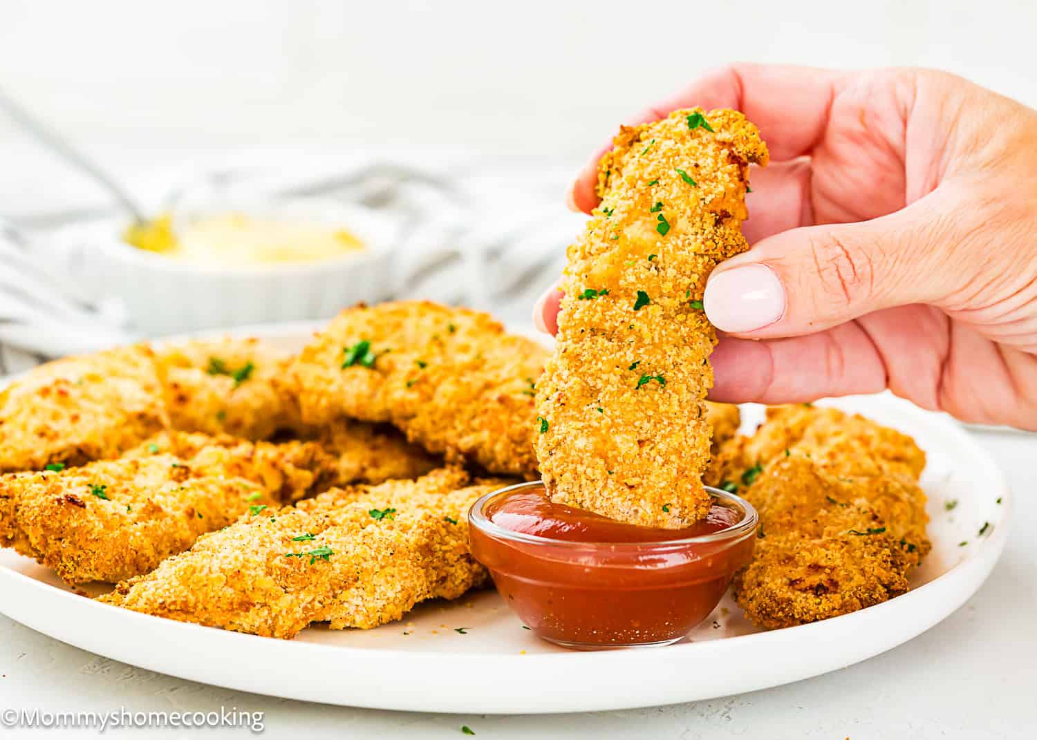 A hand dips a Homemade Chicken Tender (Egg & Dairy-Free) into a small bowl of sauce, with more chicken tenders arranged on a white plate in the background.