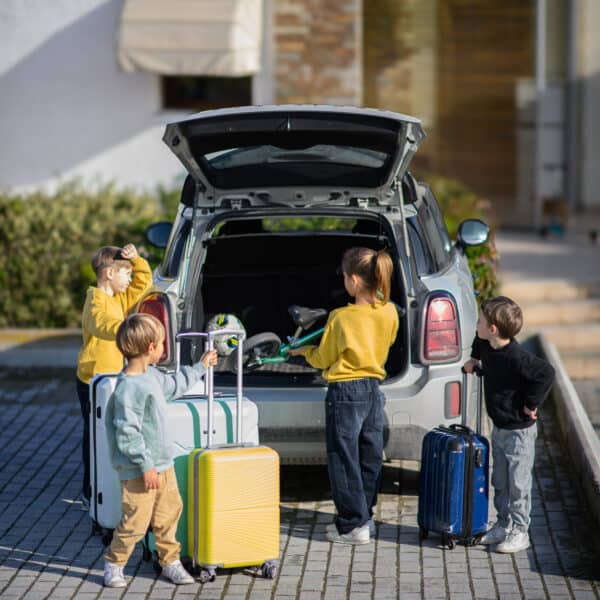 Four children stand near a parked car with the trunk open, each holding or standing by suitcases, preparing for a trip—showing how we travel with food allergies (and still have fun!) as a family.