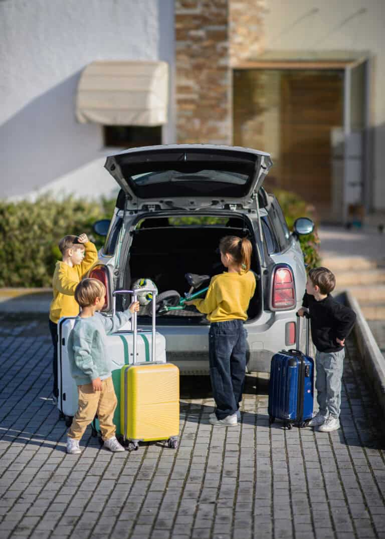 Four children stand near a parked car with the trunk open, each holding or standing by suitcases, preparing for a trip—showing how we travel with food allergies (and still have fun!) as a family.