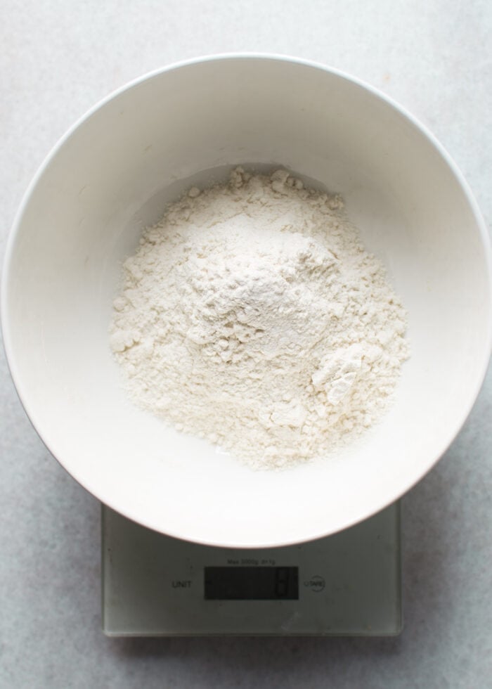 A white bowl filled with flour is placed on a digital kitchen scale, showing a key step in How-to-make-my-recipes-gluten-free by measuring alternative flours accurately.