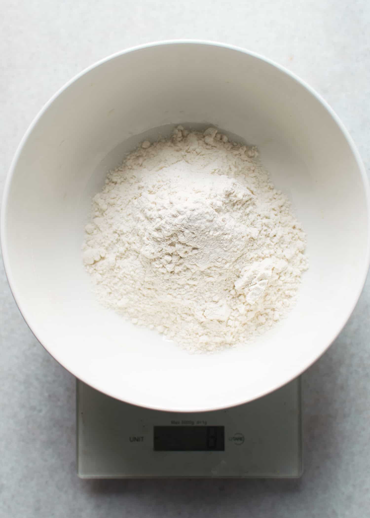A white bowl filled with flour is placed on a digital kitchen scale, showing a key step in How to make my recipes gluten-free by measuring alternative flours accurately.