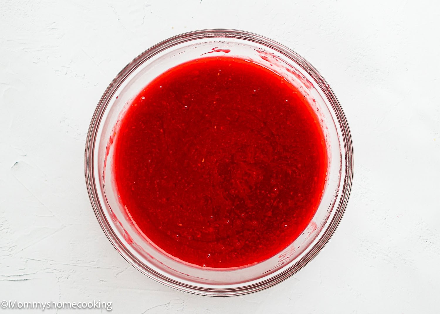 A clear glass bowl filled with bright red raspberry puree sits on a white surface, perfect for trying alongside a Raspberry Dessert Sauce Recipe.