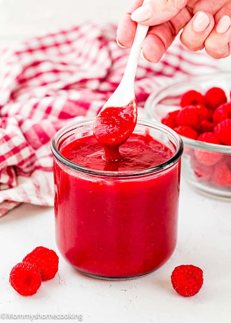 A hand holds a white spoon dipping into a glass jar of raspberry sauce, with fresh raspberries and a checkered cloth in the background—perfect for trying out your favorite Raspberry Dessert Sauce Recipe.