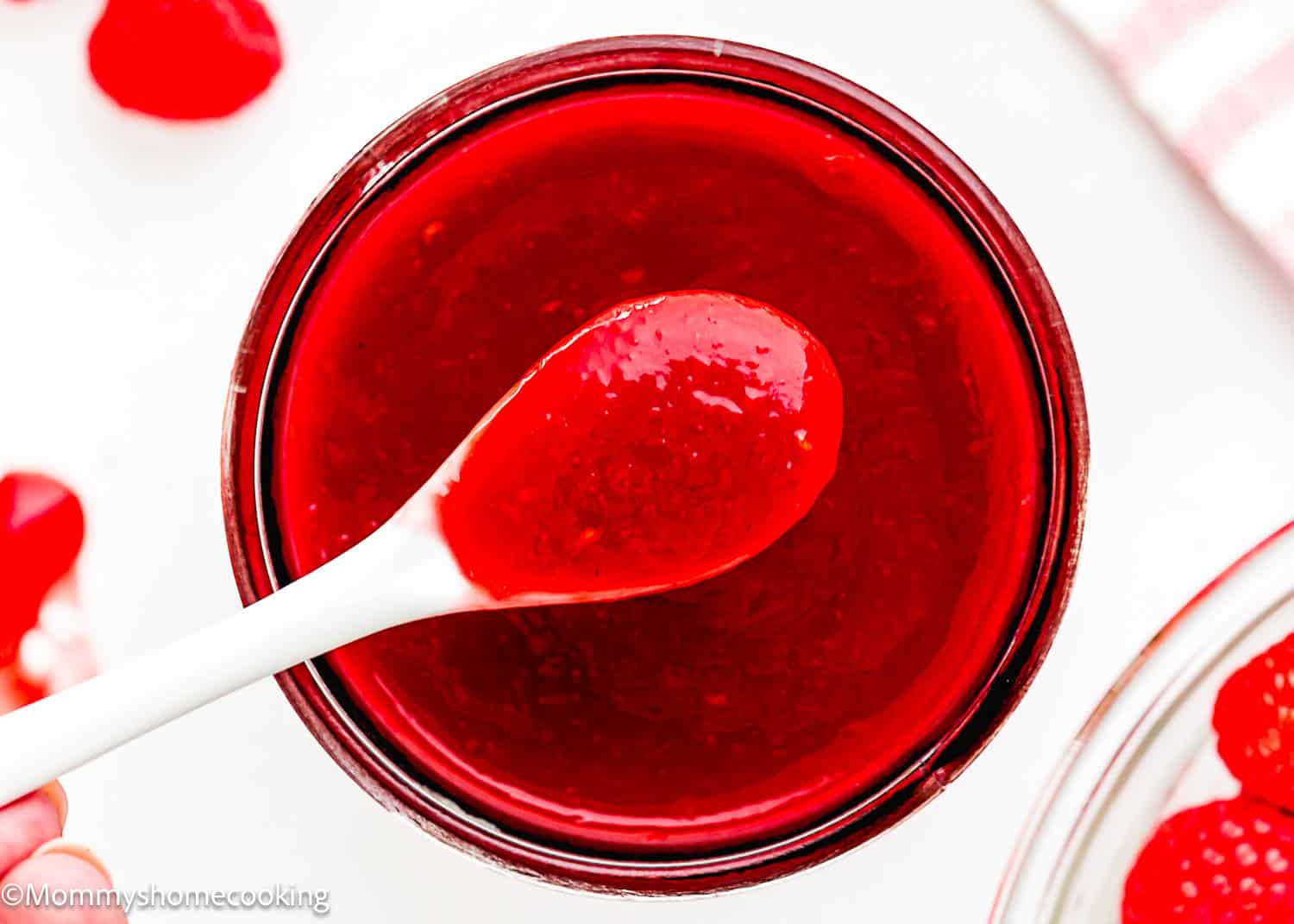 A white spoon holds a portion of glossy red raspberry dessert sauce above a glass jar filled with the same sauce.