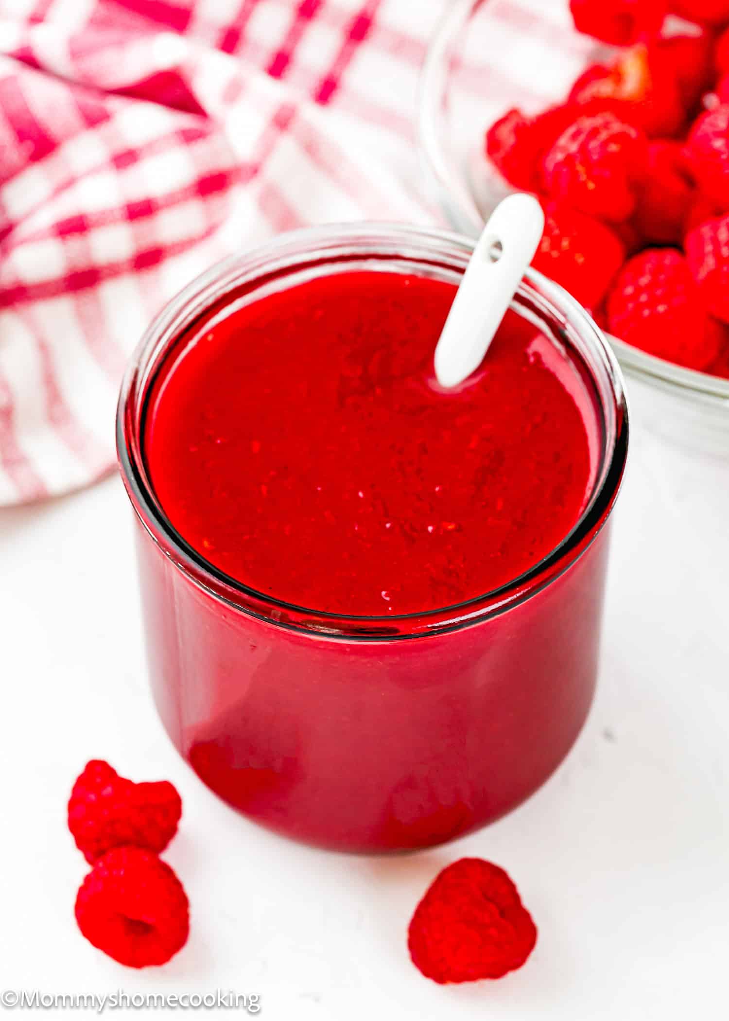 A glass jar filled with raspberry dessert sauce recipe, with a white spoon inside, sits next to fresh raspberries and a bowl of raspberries on a white surface.