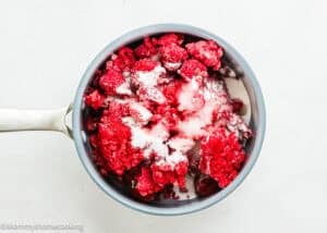 A saucepan filled with frozen raspberries and sugar on a white surface, ready to be transformed into a delicious Raspberry Dessert Sauce Recipe.