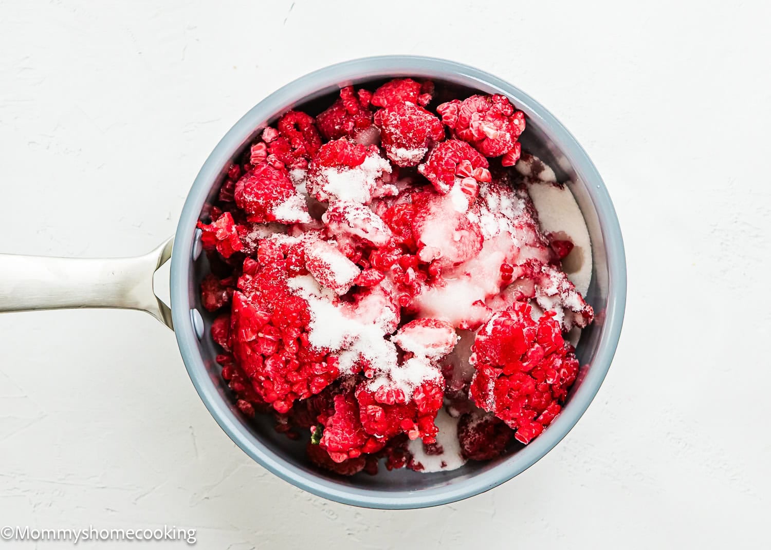 A saucepan filled with frozen raspberries and sugar on a white surface, ready to be transformed into a delicious Raspberry Dessert Sauce Recipe.