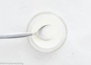 A glass bowl filled with a cornstarch slurry, with a metal spoon resting inside on a white surface—perfect for pairing with Raspberry Dessert Sauce Recipe.