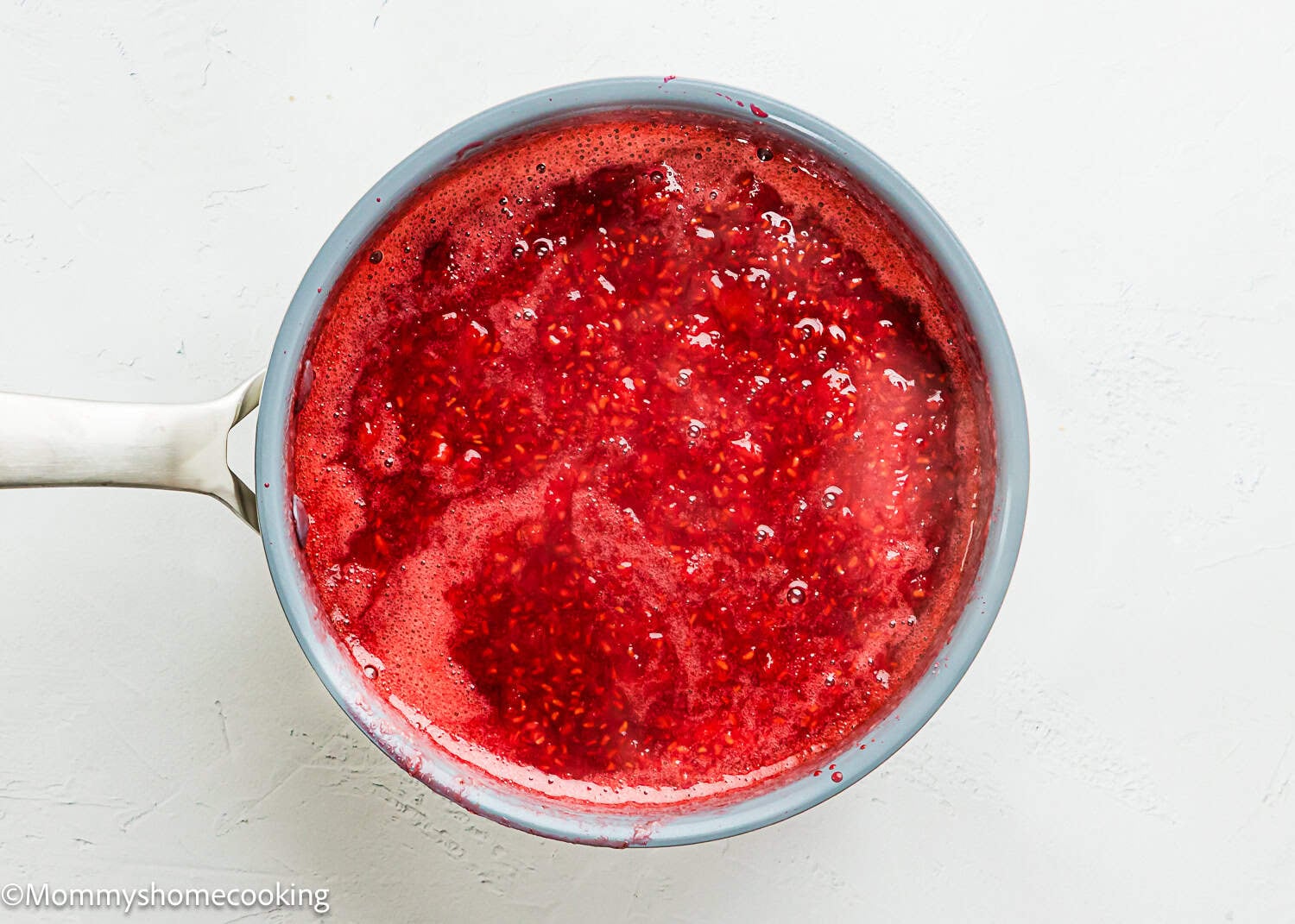 A saucepan filled with bubbling red raspberry sauce sits on a white surface, perfect for trying a new Raspberry Dessert Sauce Recipe.