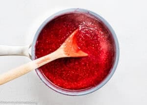 A saucepan filled with bright red Raspberry Dessert Sauce Recipe is being stirred with a wooden spoon on a white surface.