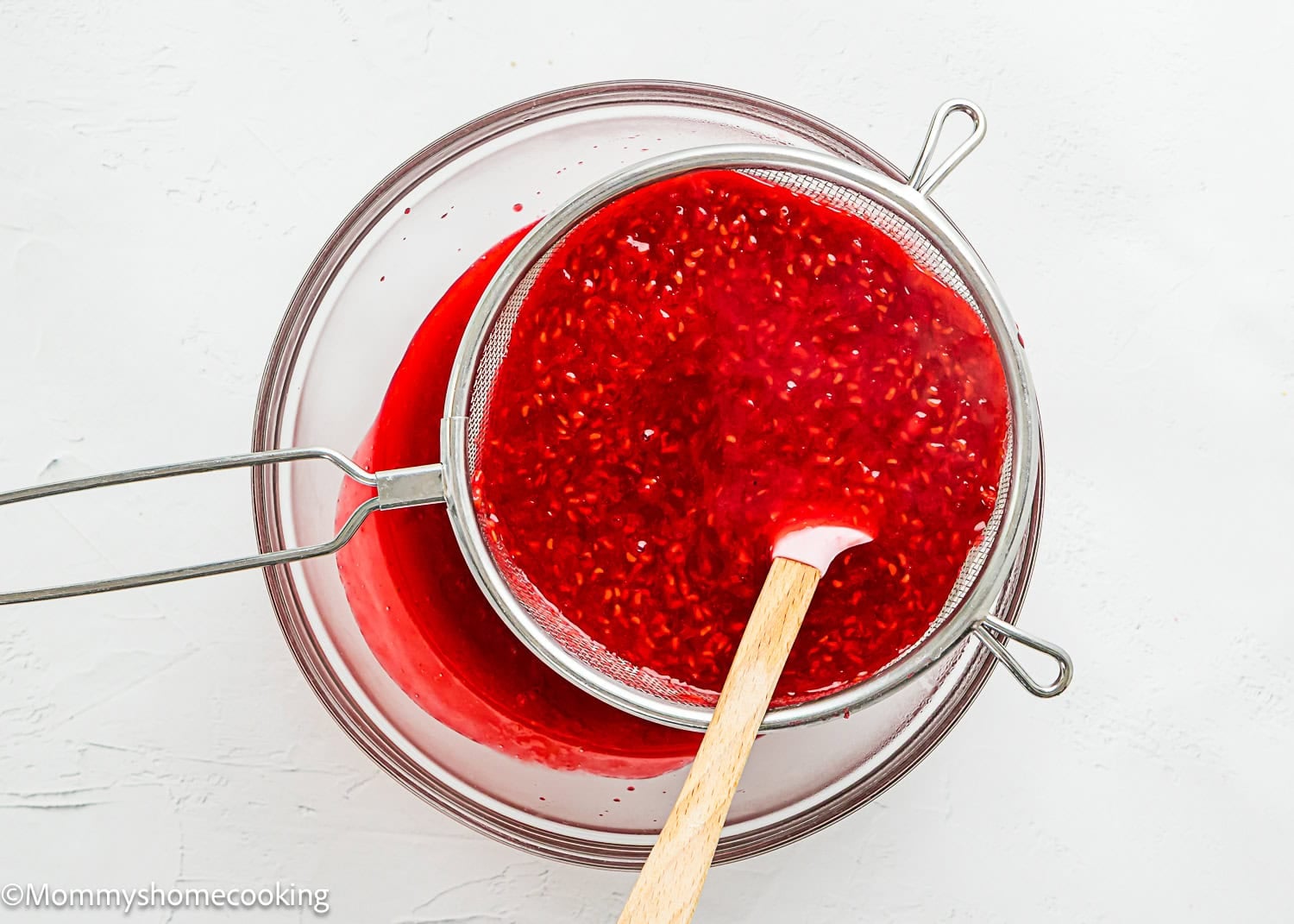 A metal strainer with raspberry sauce and seeds sits over a glass bowl, as a red spatula presses the mixture for this Raspberry Dessert Sauce Recipe.