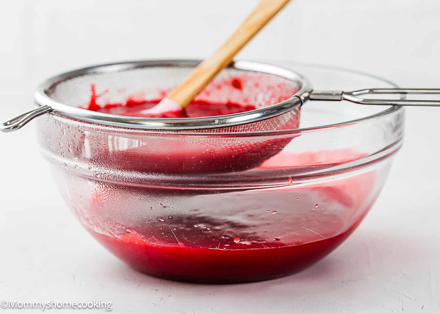 A glass bowl with red fruit puree being strained through a fine mesh sieve using a wooden spatula, perfect for preparing a Raspberry Dessert Sauce Recipe.