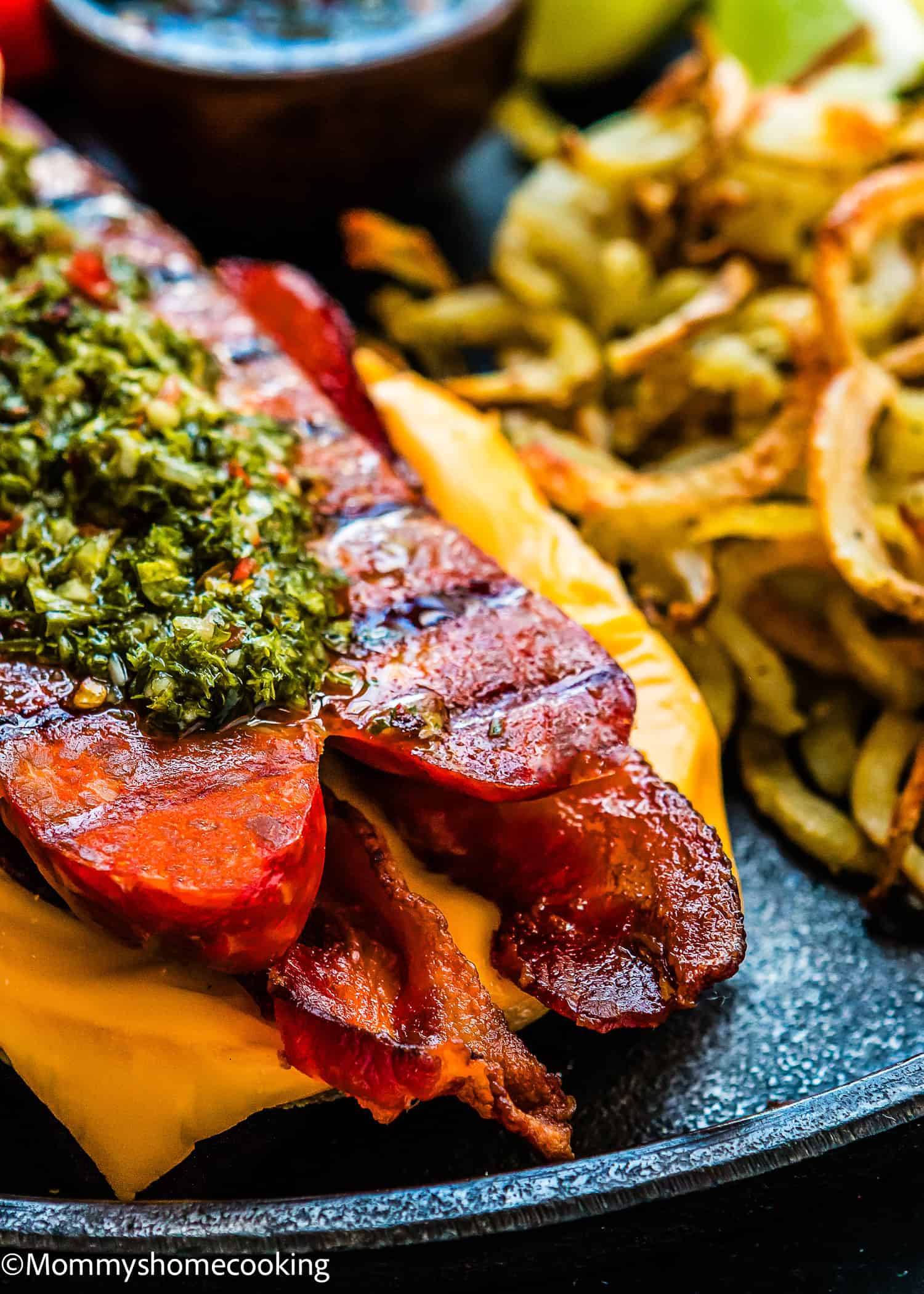 Easy Choripan with Chimichurri Sauce: Grilled sausage topped with chimichurri, served with bacon, melted cheese, and crispy fries on a black plate.