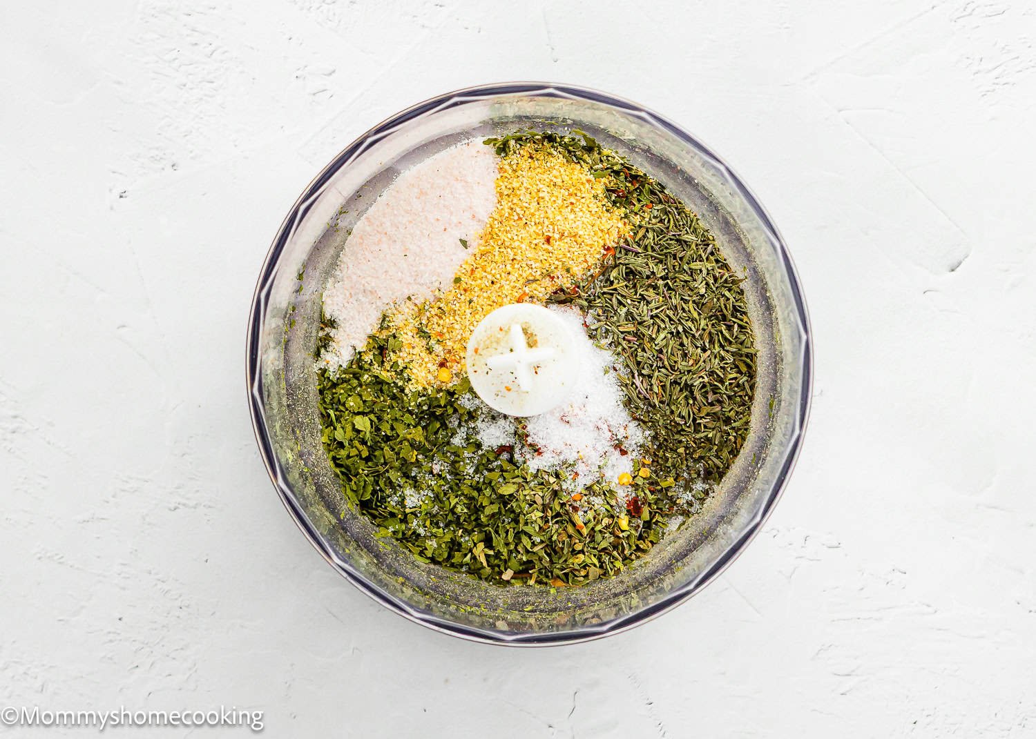 A food processor bowl containing separate piles of spices and dried herbs on a white surface, ready to be mixed into an Easy Dry Chimichurri Seasoning (Allergy-Safe & Full of Flavor!).