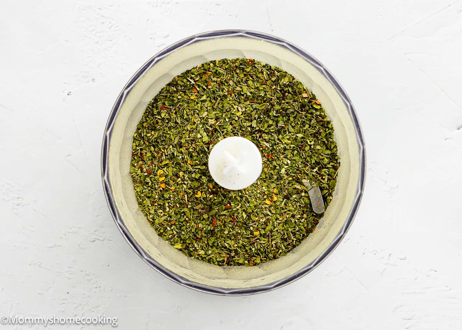 A food processor bowl filled with chopped green herbs and spices for Easy Dry Chimichurri Seasoning (Allergy-Safe & Full of Flavor!), viewed from above on a white surface.