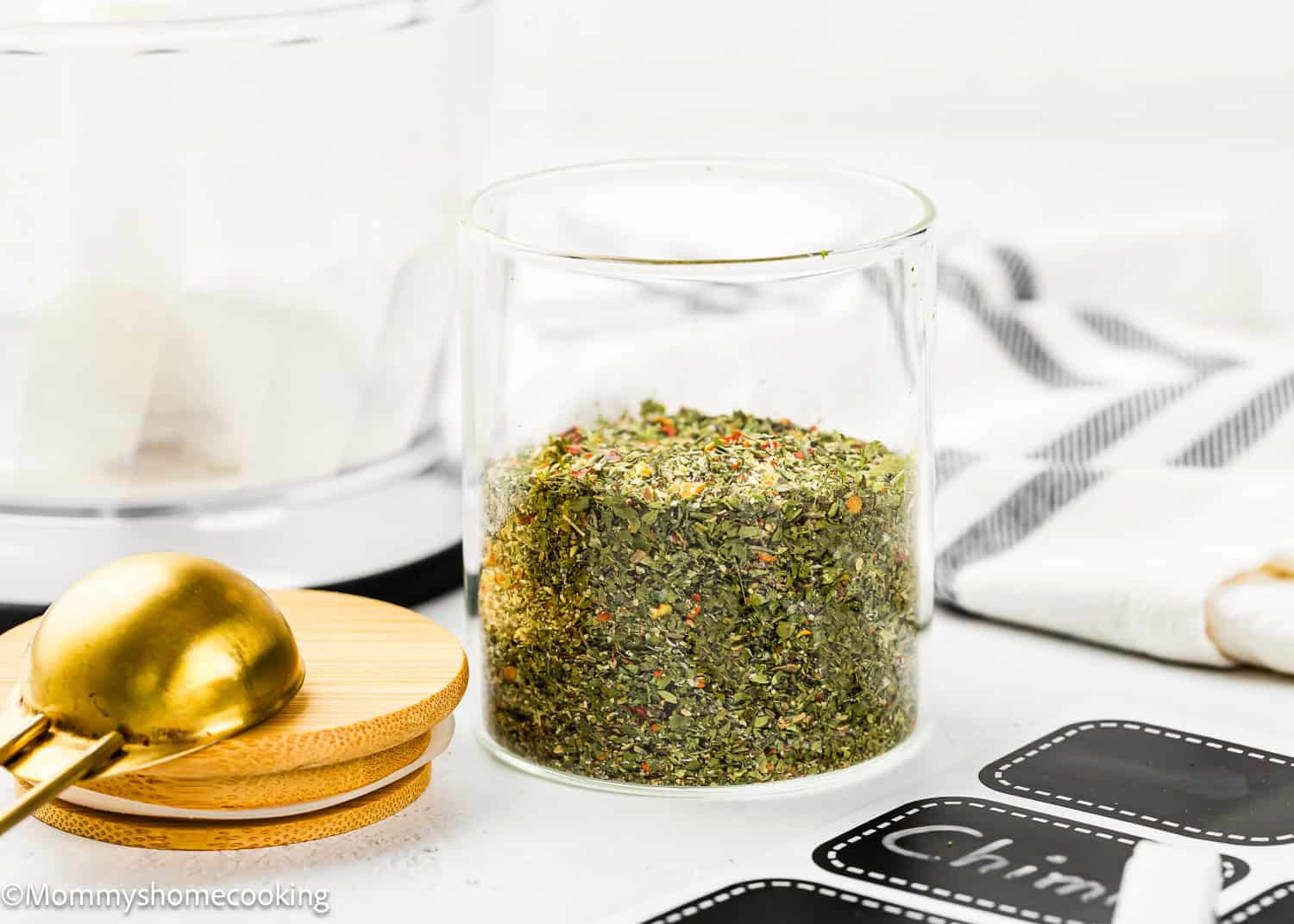 A glass jar filled with Easy Dry Chimichurri Seasoning (Allergy-Safe & Full of Flavor!) sits on a white surface beside a gold measuring spoon and blank black labels.