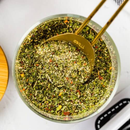 A gold measuring spoon rests in a glass bowl filled with Easy Dry Chimichurri Seasoning (Allergy-Safe & Full of Flavor!), showcasing a vibrant mixed dried herb blend.