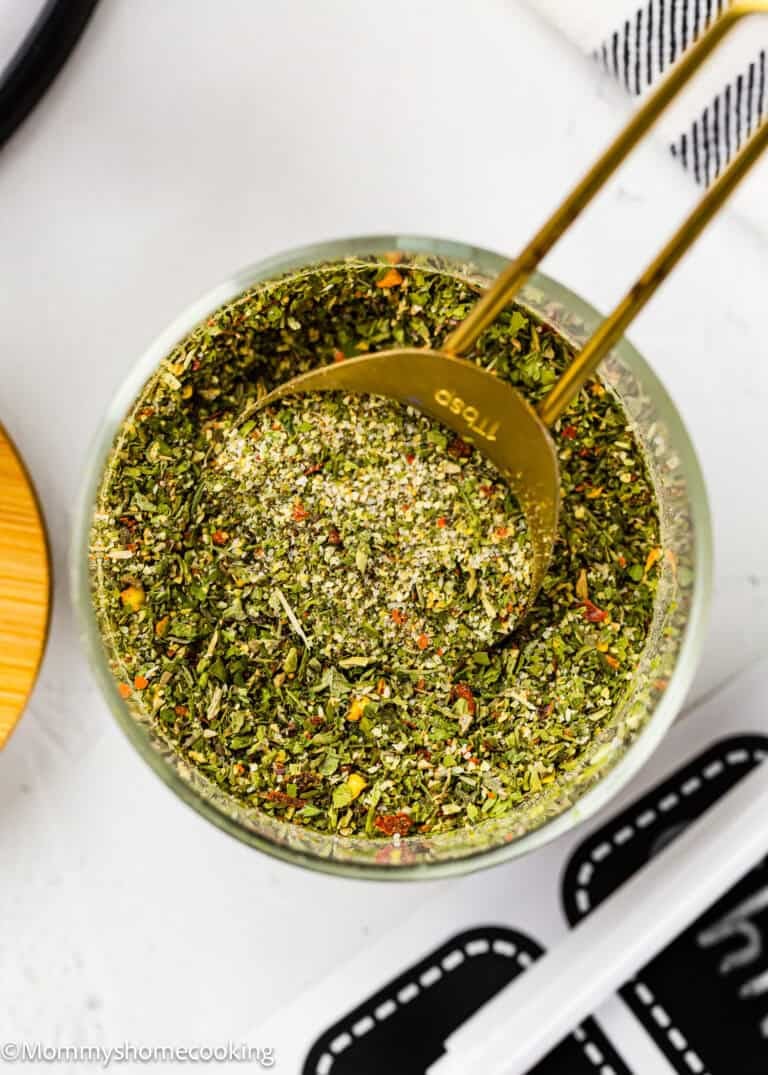 A gold measuring spoon rests in a glass bowl filled with Easy Dry Chimichurri Seasoning (Allergy-Safe & Full of Flavor!), showcasing a vibrant mixed dried herb blend.