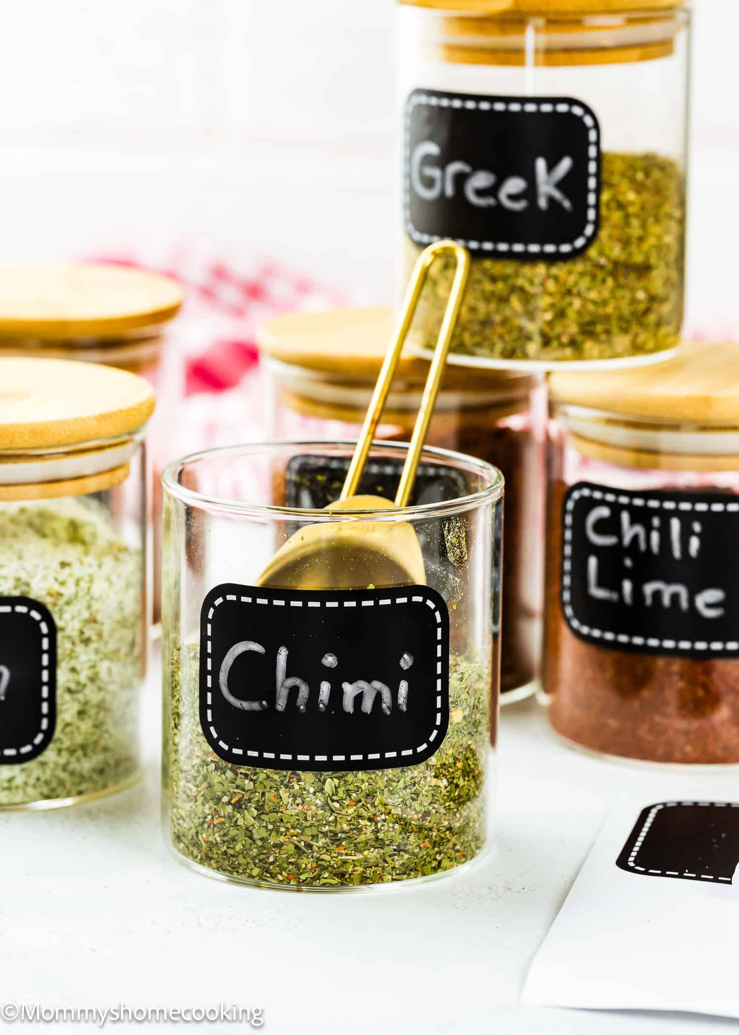 Clear glass jars with wooden lids contain various spice blends, each labeled with a black sticker, including "Chimi," featuring Easy Dry Chimichurri Seasoning (Allergy-Safe & Full of Flavor!), "Greek," and "Chili Lime." A gold spoon rests in the Chimi jar.