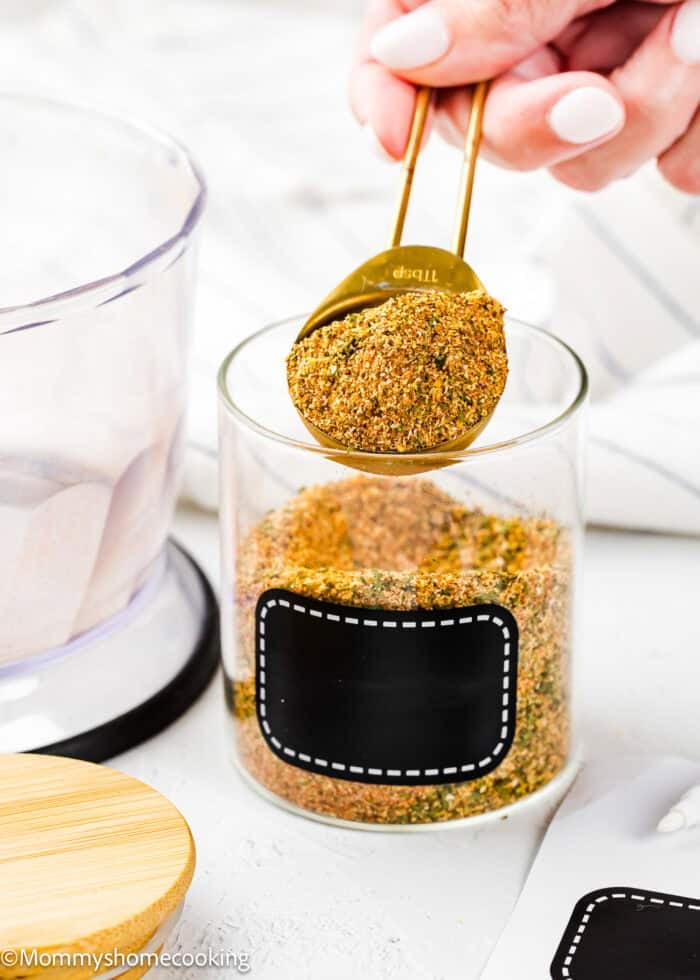 A hand holds a tablespoon of Easy Homemade Everyday Seasoning (All-Purpose & Allergy-Safe) over a clear glass jar filled with the same blend; the jar has a blank black label.